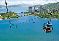 A wide view of a bicable gondola lift on Lantau Island in Hong Kong, surrounded by a large body of water below, as well as a city scene.