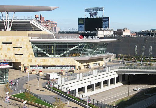 A wide exterior view of the Target Field, with cars, a scoreboard, flags, a parking lot, and building structures in view.